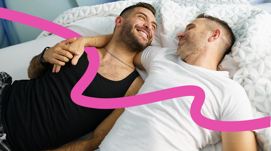 Two men cuddle in bed, smiling and enjoying a warm moment together.
