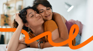 Two women hug on a sofa, smiling softly as they share a tender moment, with an orange decorative word across the foreground.