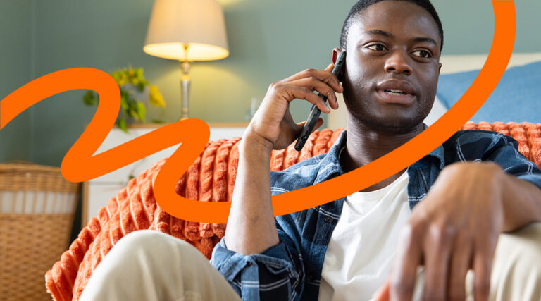 A young man sits on a couch talking on a cellphone in a cozy living room, with a warm lamp and orange knit blanket nearby.